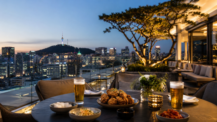 루프탑 테이블 위에 치킨과 맥주가 놓이고 서울 도심 풍경이 배경으로 보이는 장면