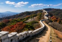 The Seoul City Wall winds along a mountain ridge with autumn trees and N Seoul Tower in the distance